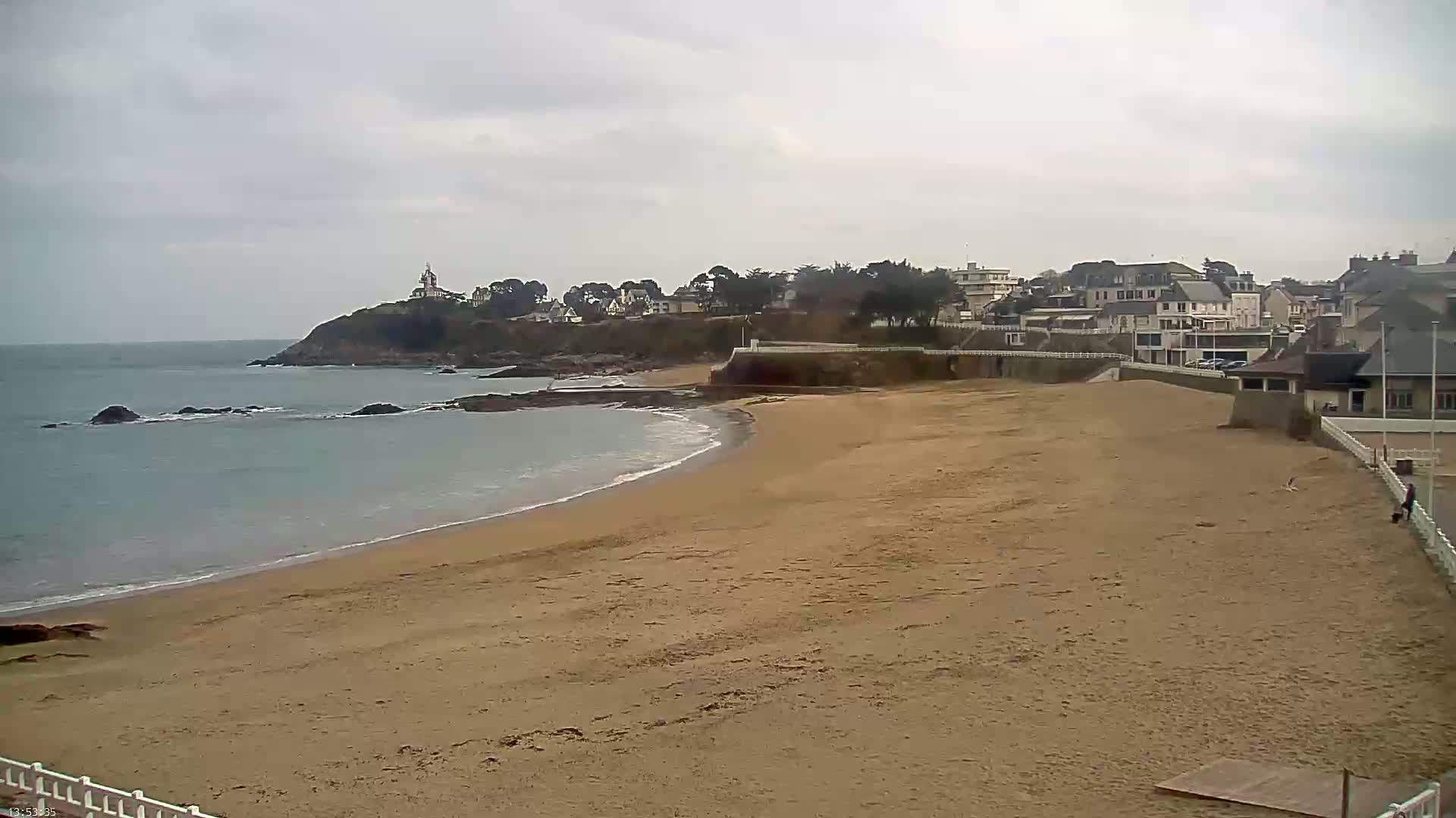 A sandy beach curves gently into a calm ocean under an overcast sky, with a town and a small lighthouse visible on a distant headland.