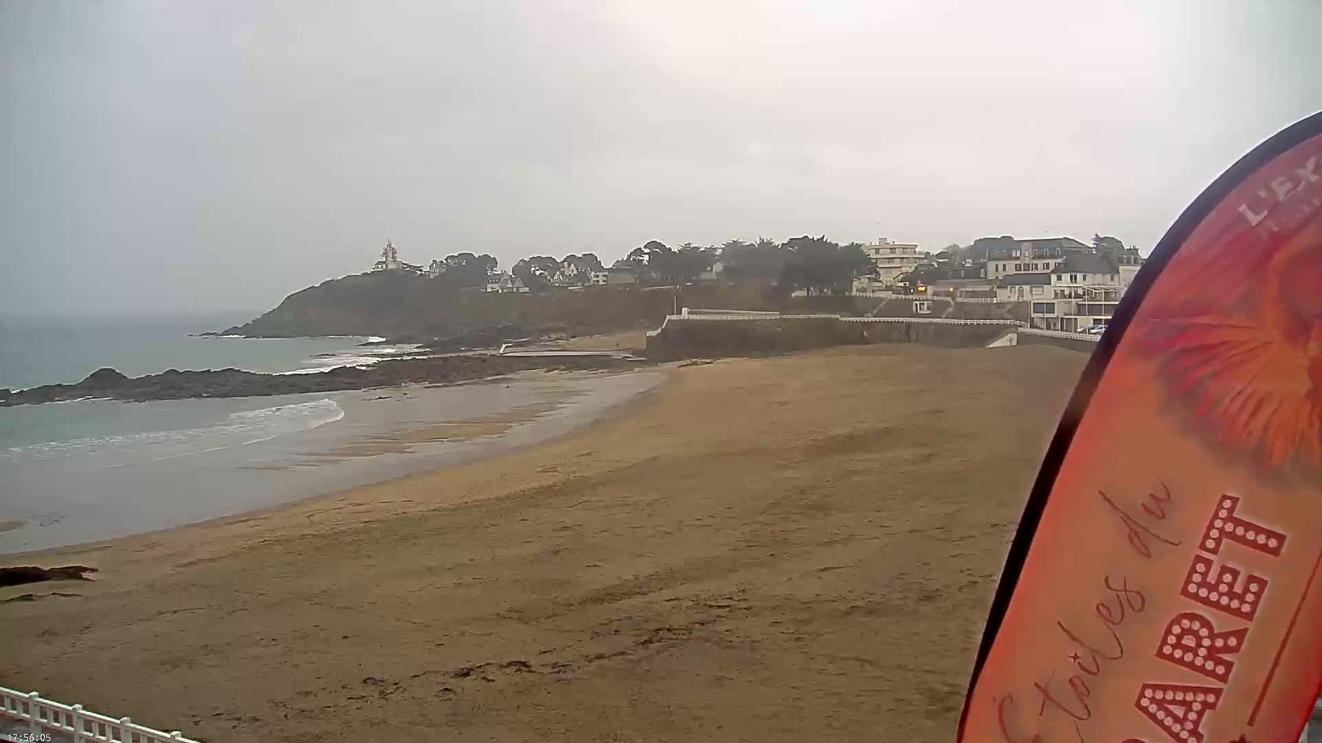 A sandy beach stretches out to a calm ocean under an overcast sky, with a small town and a lighthouse visible on a nearby cliff.