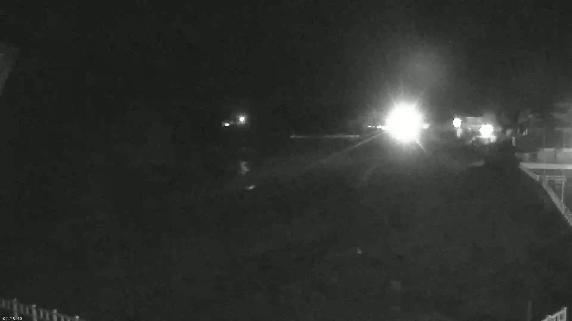 A nighttime, low-light image shows a road with headlights from approaching vehicles, and a partially visible structure to the right.