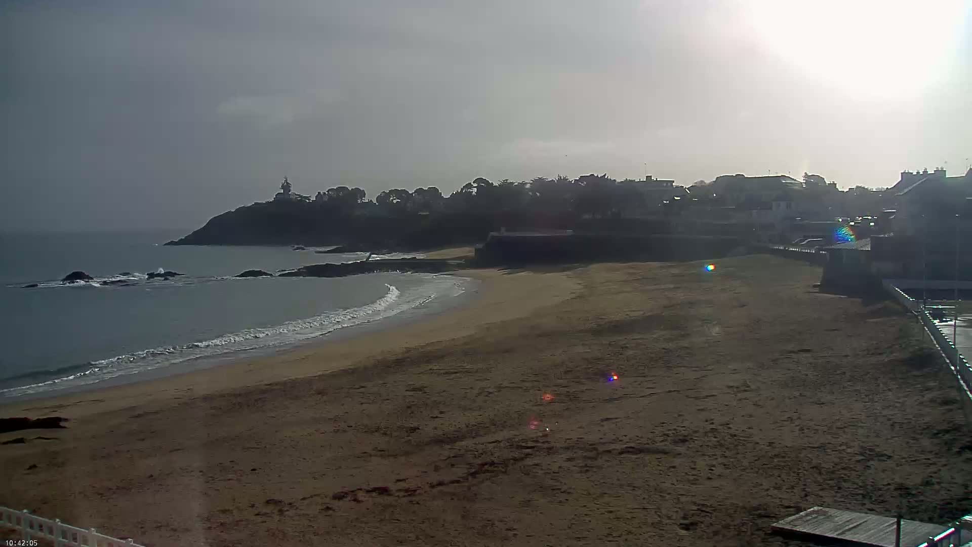 A sandy beach curves gently to the left, meeting a calm ocean with small waves;  a hazy sun shines brightly in an overcast sky above a distant coastline with buildings and a small lighthouse.