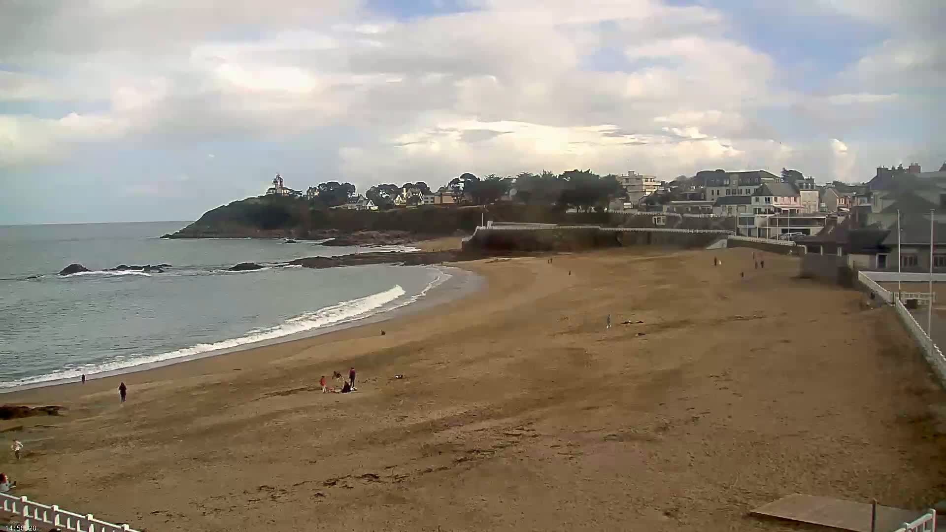 A sandy beach with a few people on it curves around a rocky point where houses and a lighthouse are visible under a partly cloudy sky.