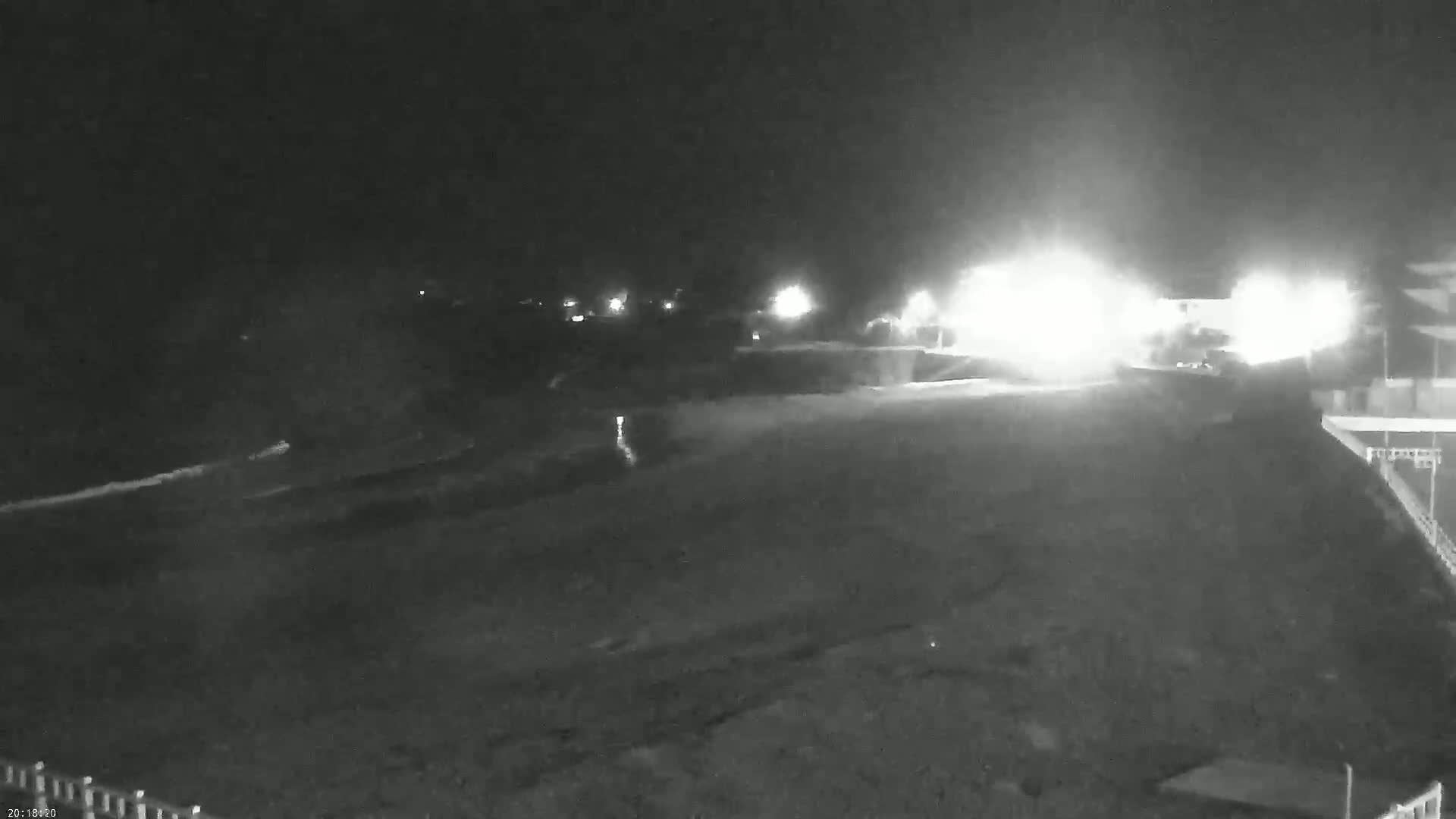 A nighttime, low-light image shows a dark, mostly empty beach or shoreline with bright lights in the distance and faint waves visible near the bottom.