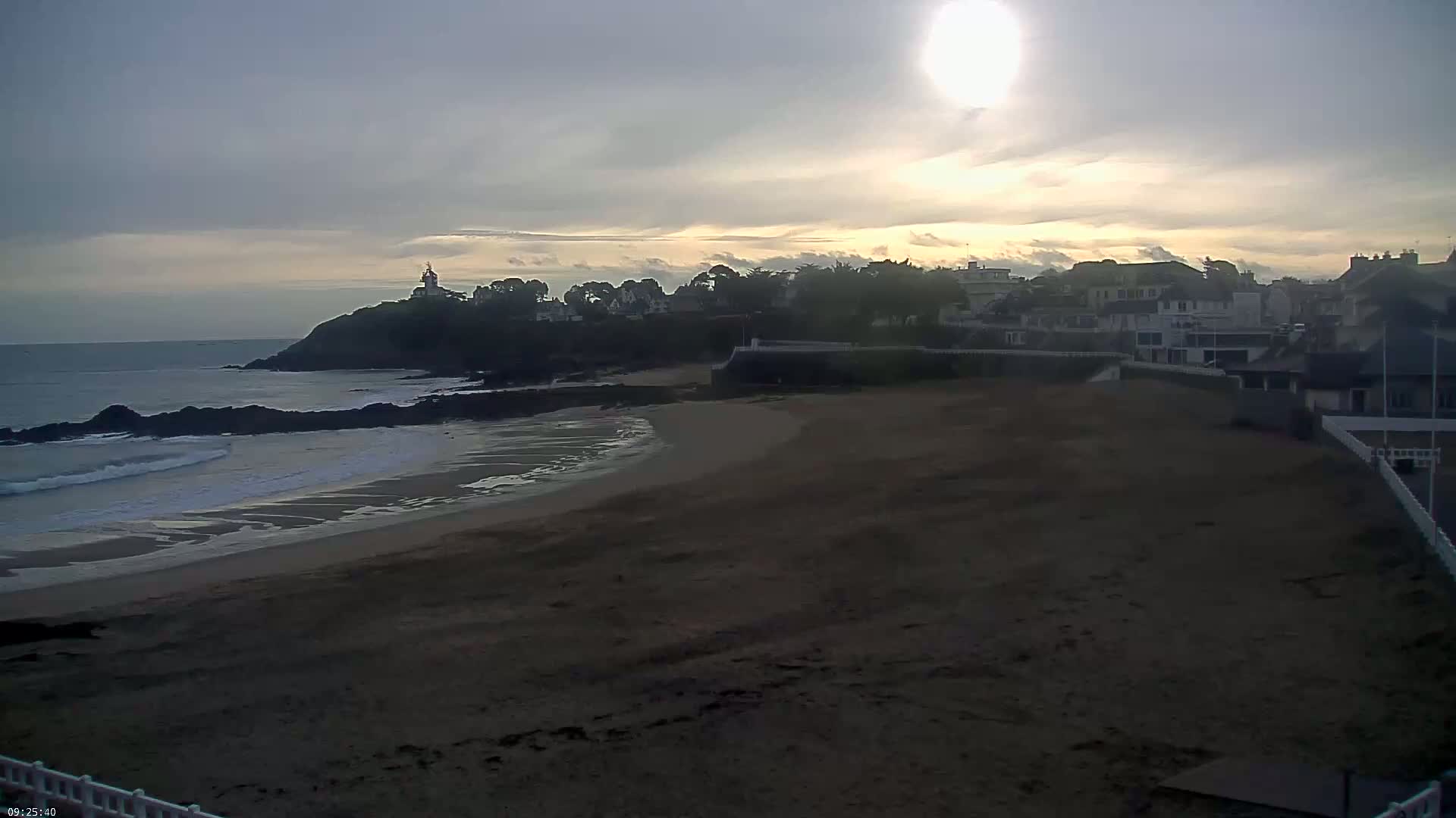 A sandy beach stretches out toward a calm ocean under a mostly cloudy sky, with a small town and lighthouse visible on a distant headland.