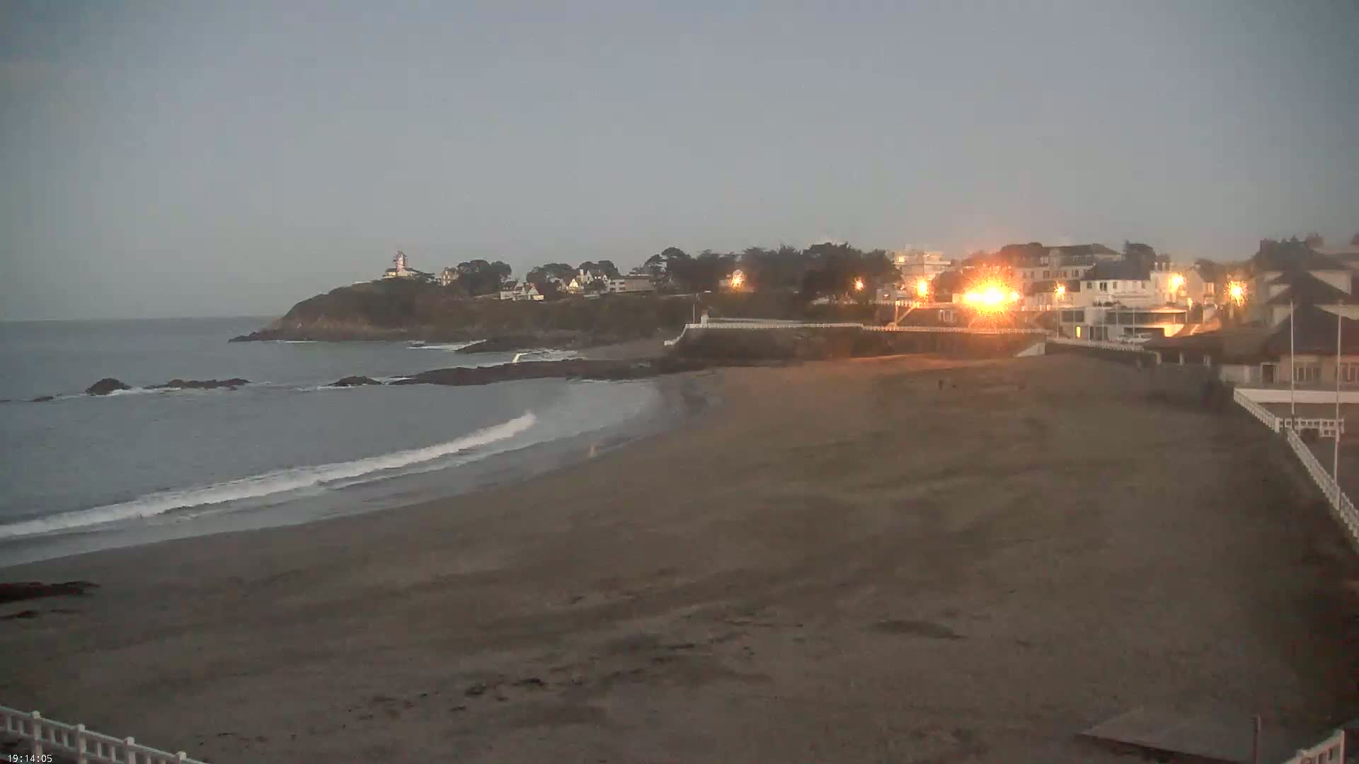 A sandy beach at dusk shows small waves lapping the shore, with a small town and lighthouse visible on a cliff in the distance.