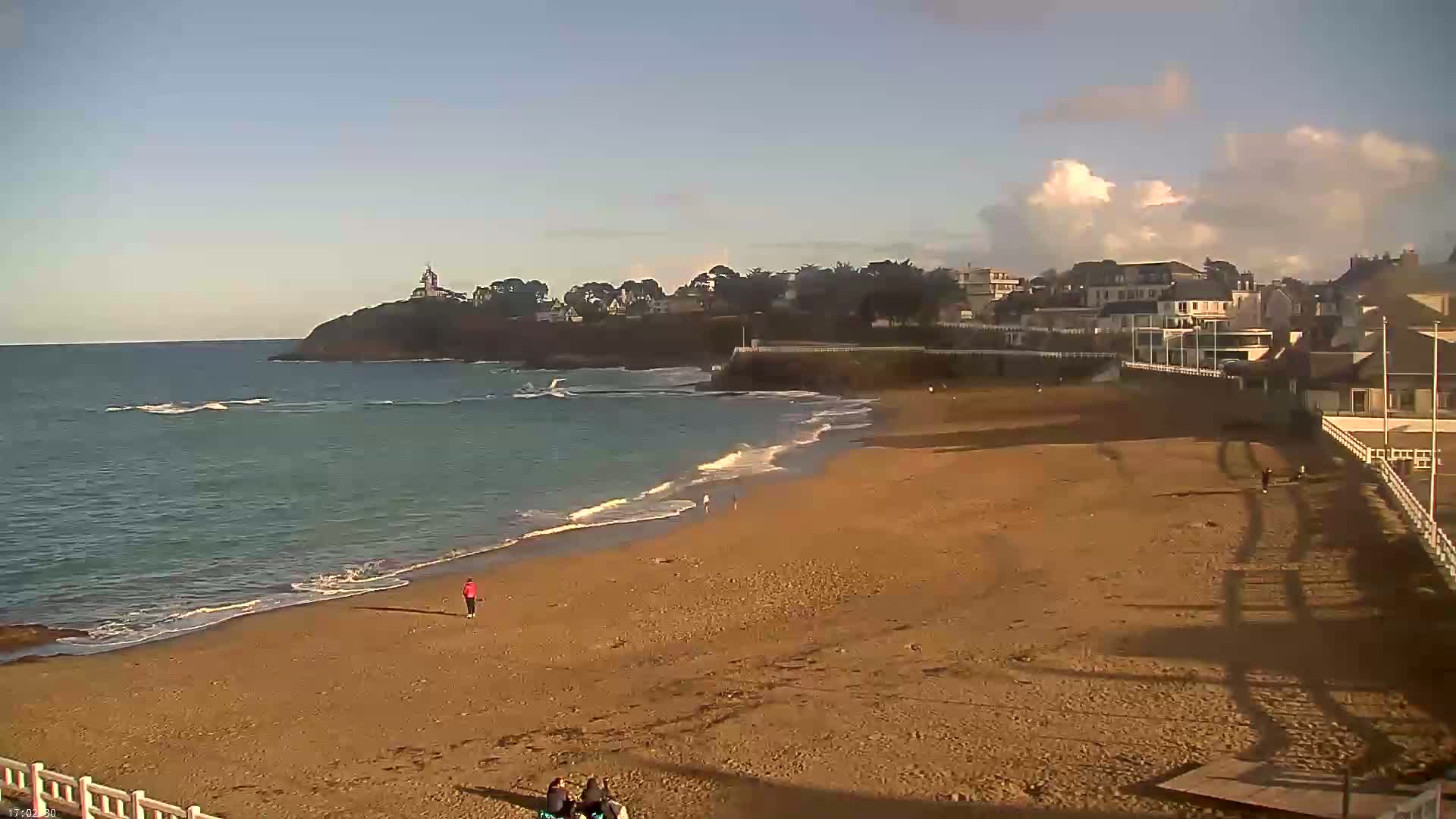 A sandy beach stretches out to a calm sea under a partly cloudy sky, with a few people walking along the shore and buildings visible on a distant headland.