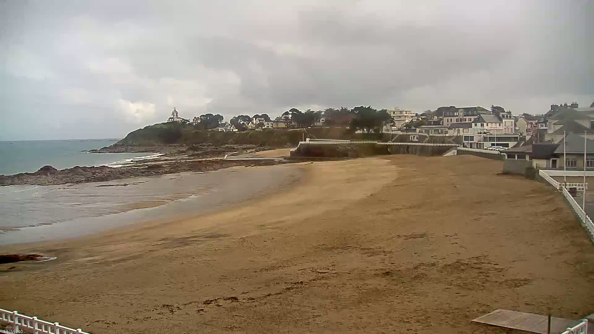 A sandy beach stretches out to a calm ocean under a cloudy sky, with a small town and a rocky headland visible in the background.