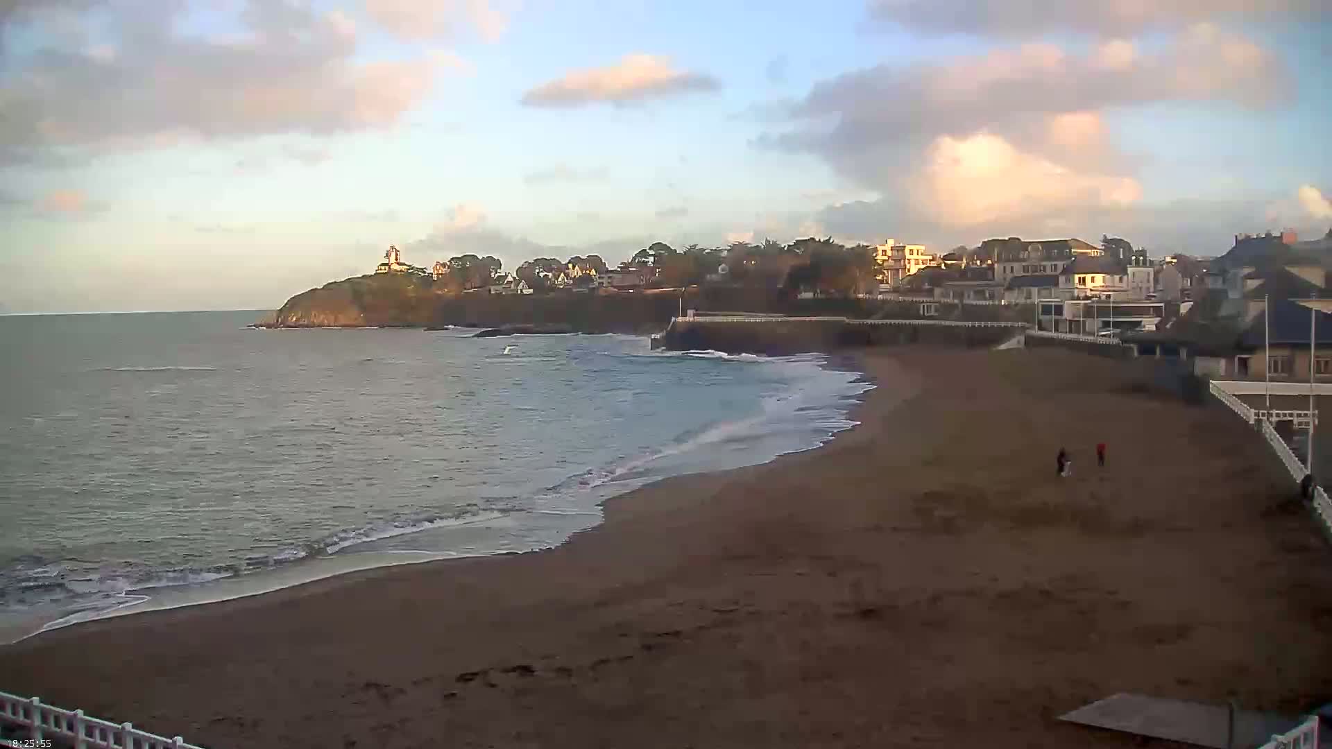 A sandy beach with gentle waves laps at the shore, under a partly cloudy sky, next to a coastal town with houses and a lighthouse on a nearby cliff.