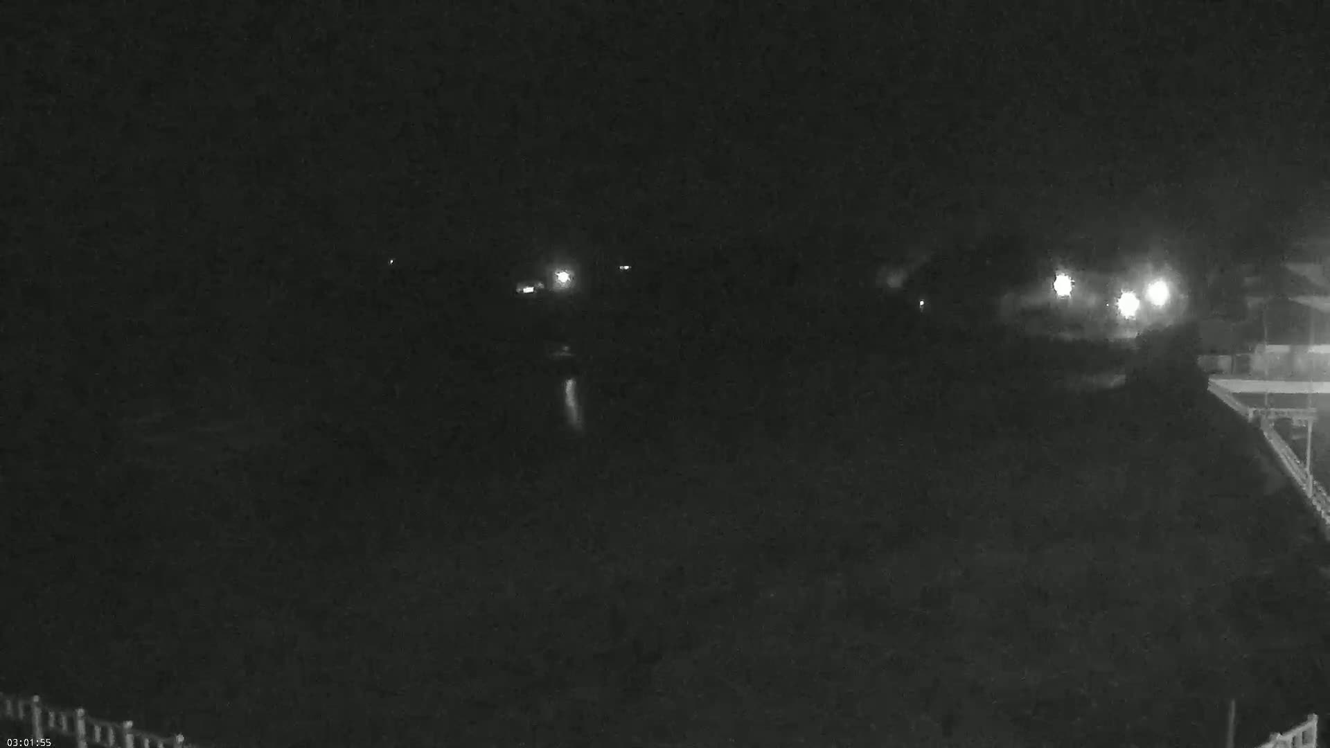 A nighttime, low-light view shows a dark body of water with a few distant lights visible on the far shore and a portion of a structure on the lower right.