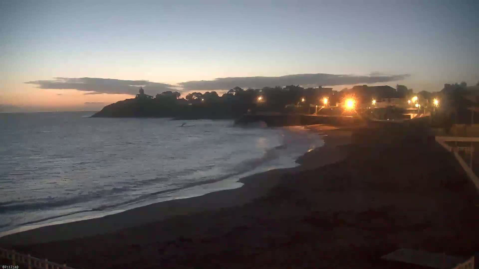A dark beach at twilight, with gentle waves lapping the shore, is illuminated by lights from buildings on a cliff overlooking the ocean.