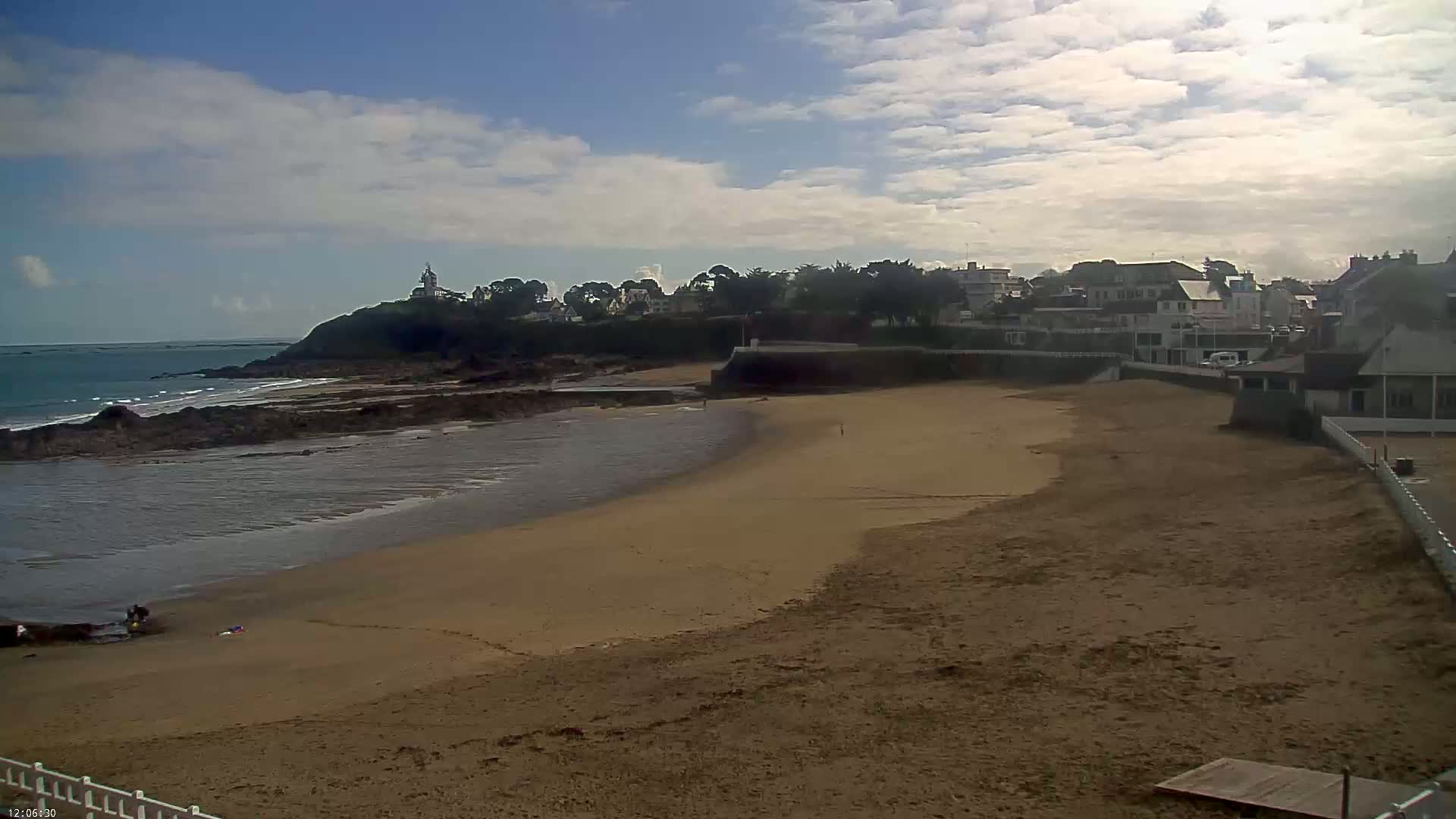 A sandy beach curves around a rocky shoreline, with a small town and a lighthouse visible on a hill in the background under a partly cloudy sky.