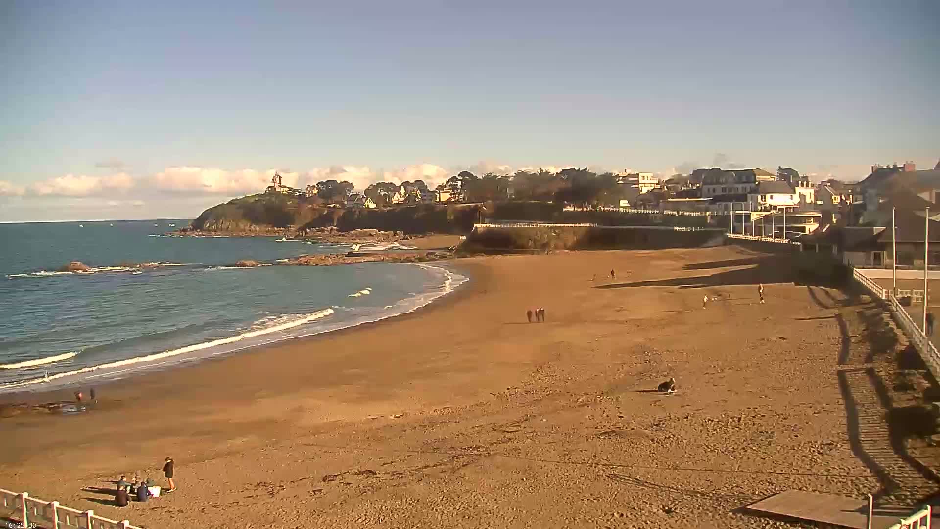 A sandy beach curves around a rocky point, with a town and lighthouse visible in the distance under a mostly clear sky.
