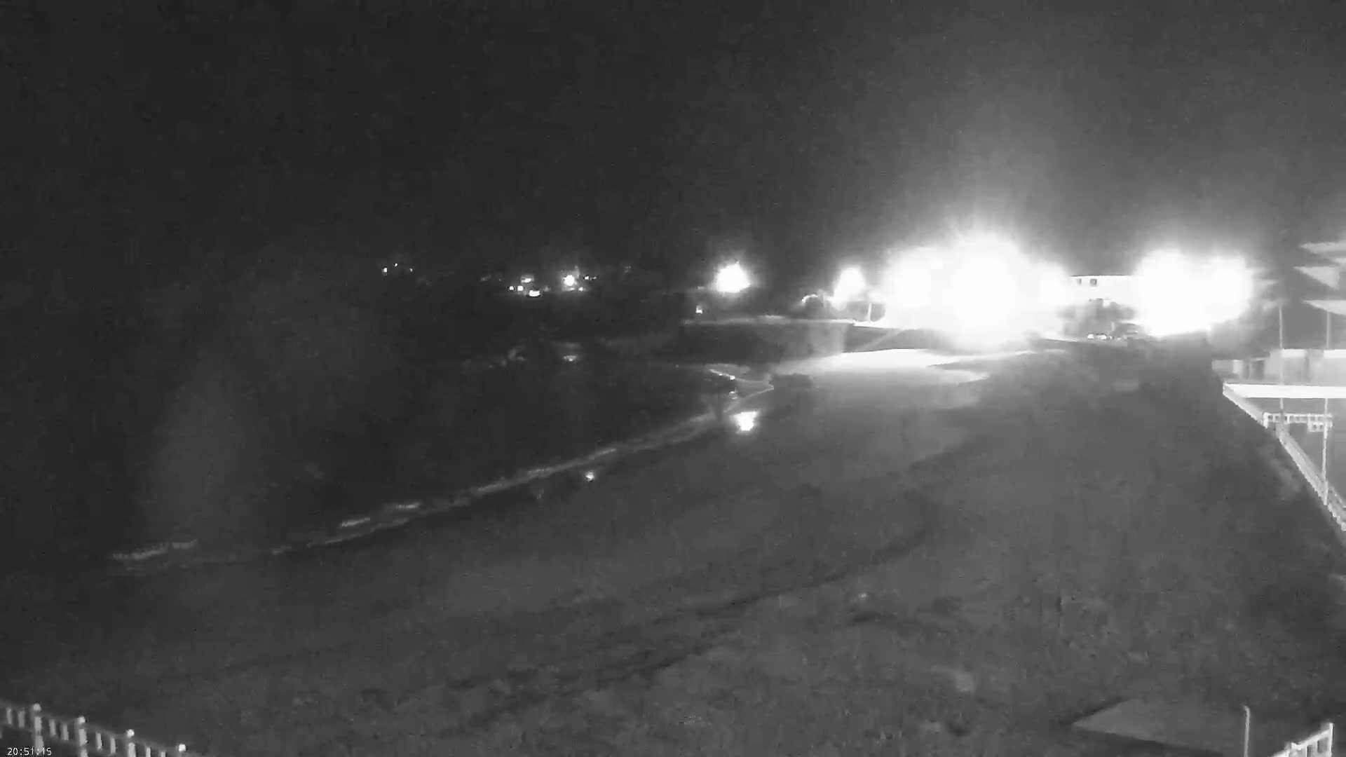 A nighttime, low-light view of a shoreline at night, showing a dark beach and water, with bright lights visible in the distance.