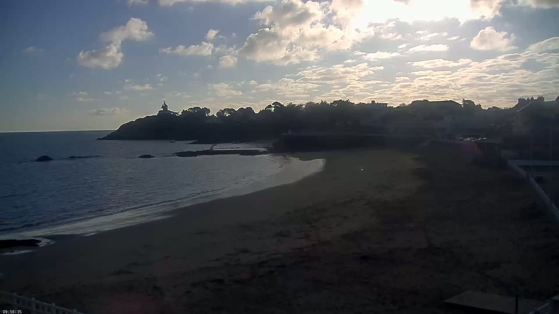 A sandy beach curves around a rocky point, where a small town sits under a partly cloudy sky, bathed in the light of a setting sun.