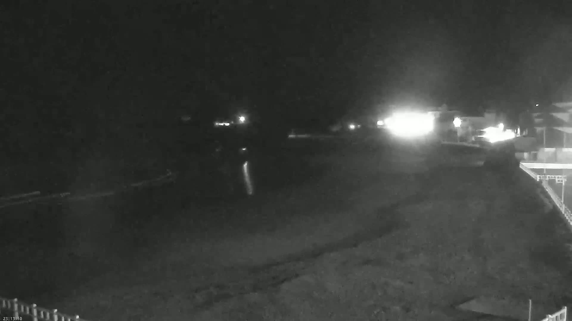 A nighttime, low-light image shows a dark, possibly sandy, area with faint lights in the distance and a building partially visible in the lower right corner.
