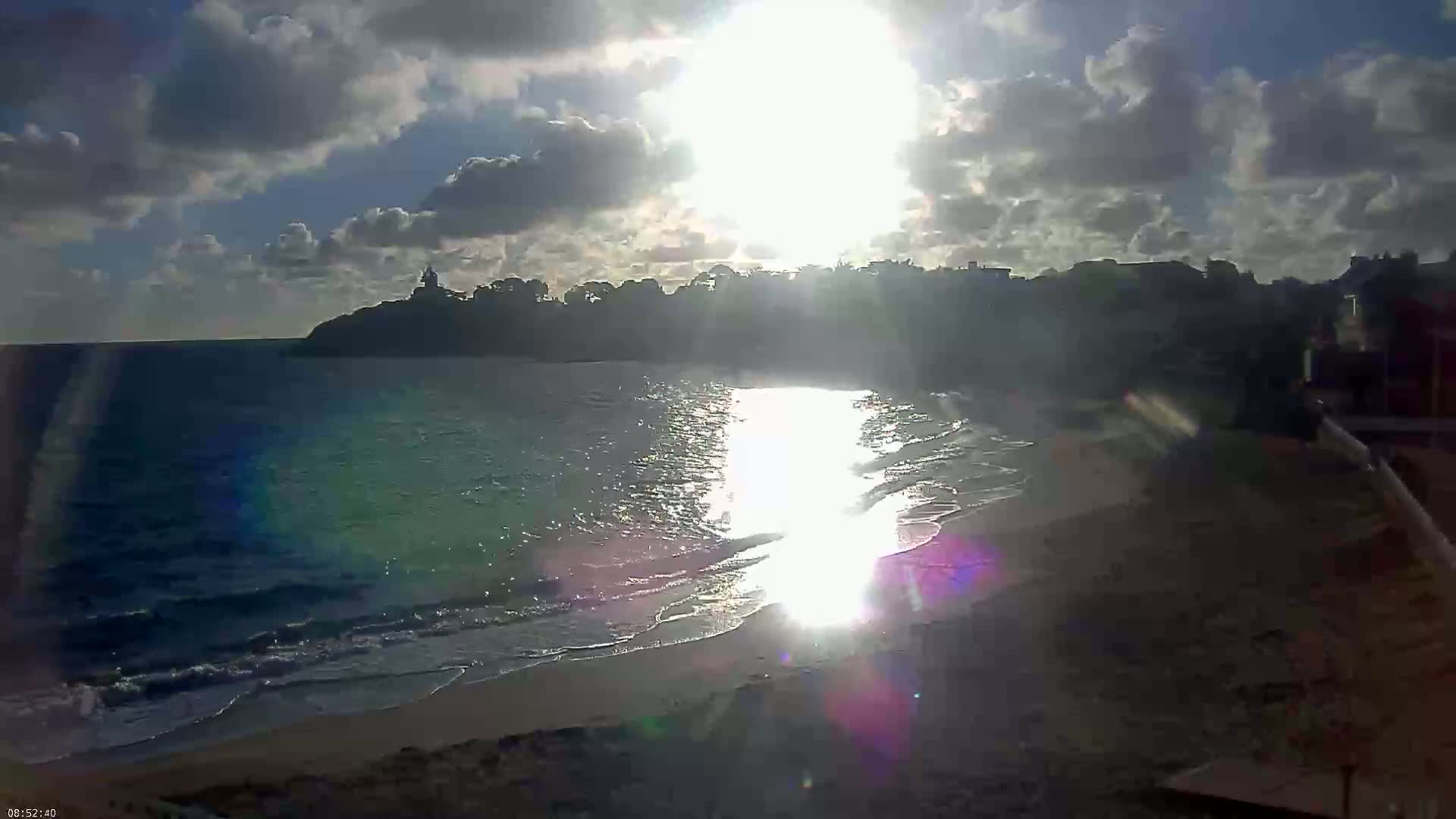 A partly cloudy sunrise shines on a sandy beach with gentle waves lapping the shore, behind which sits a dark silhouette of buildings and trees on a coastal headland.