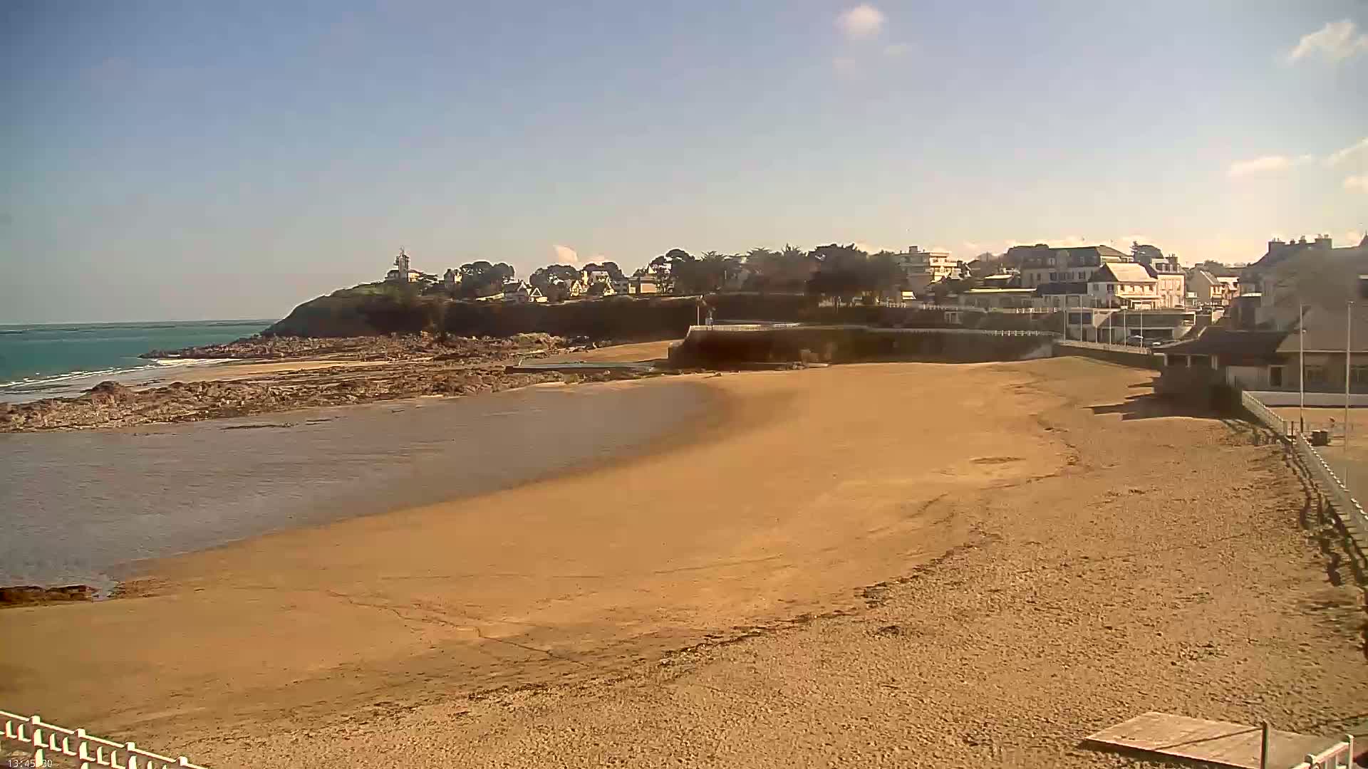 A sandy beach curves around a calm inlet of water, with a small town and a lighthouse visible on a cliff in the background under a mostly sunny sky.