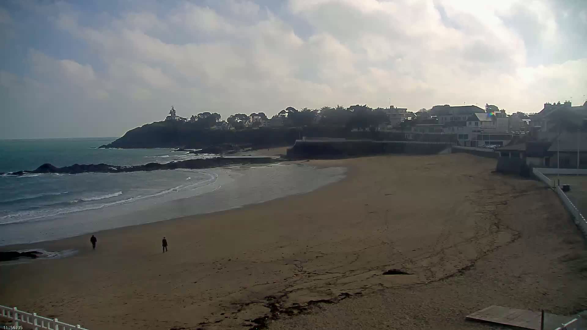 A sandy beach curves around a rocky point, with a small town visible on the cliff, under a partly cloudy sky.