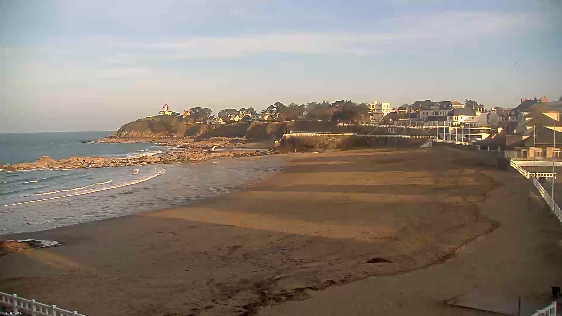 A sandy beach stretches out to a calm ocean under a mostly clear sky, with buildings and a small cliff visible in the background.