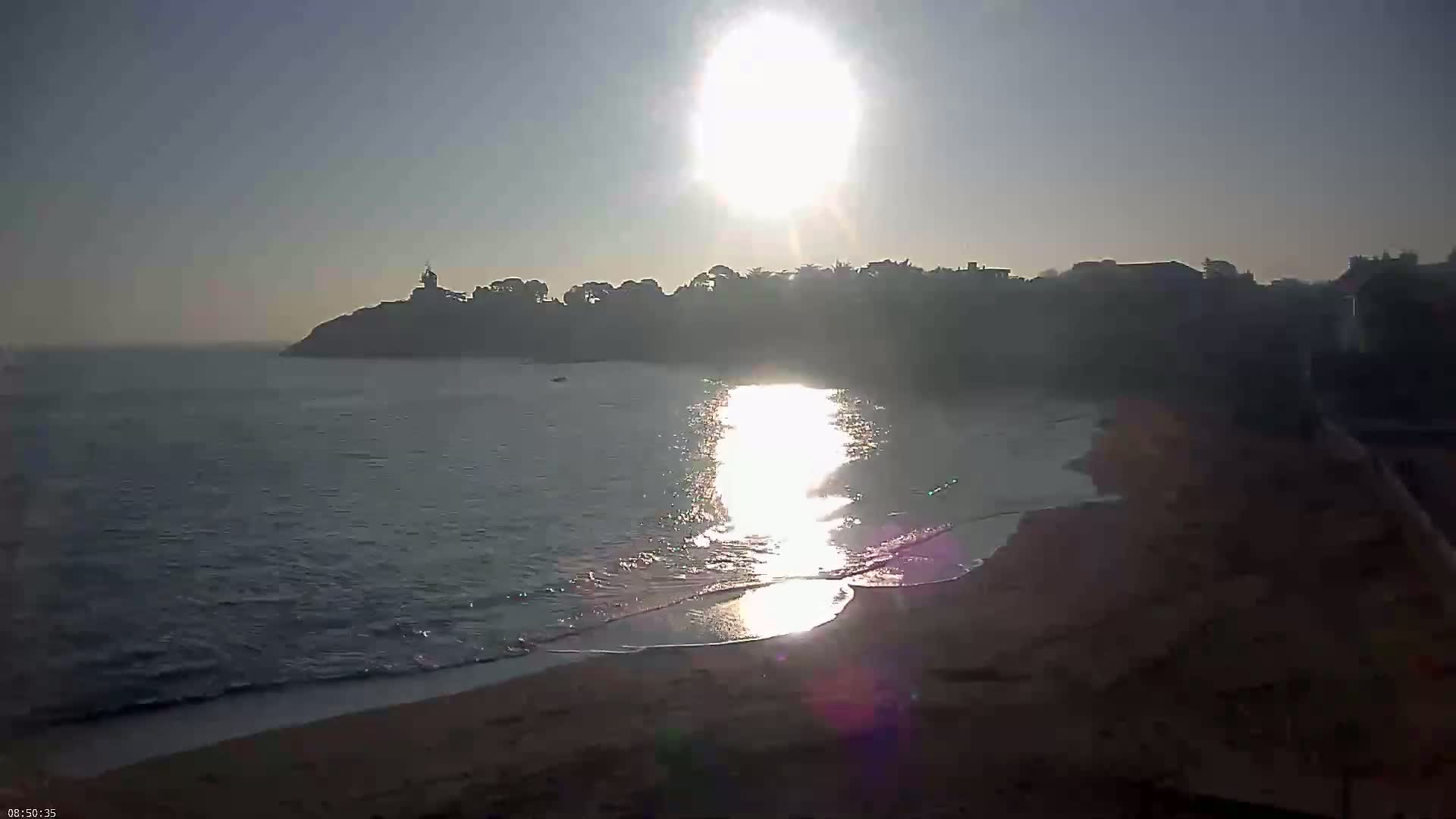 A bright sun shines over a calm ocean lapping a sandy shore, with a distant headland and buildings silhouetted against the sky.