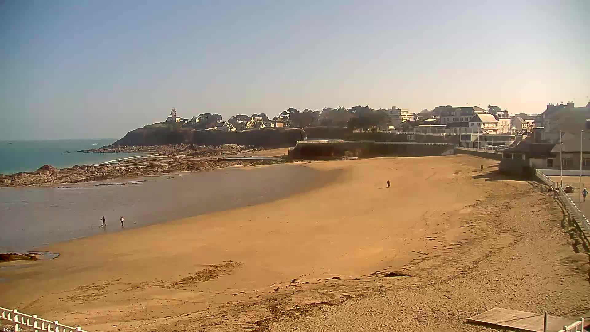 A wide sandy beach stretches toward a calm ocean, under a clear blue sky, with a small town and a lighthouse visible on a distant headland.