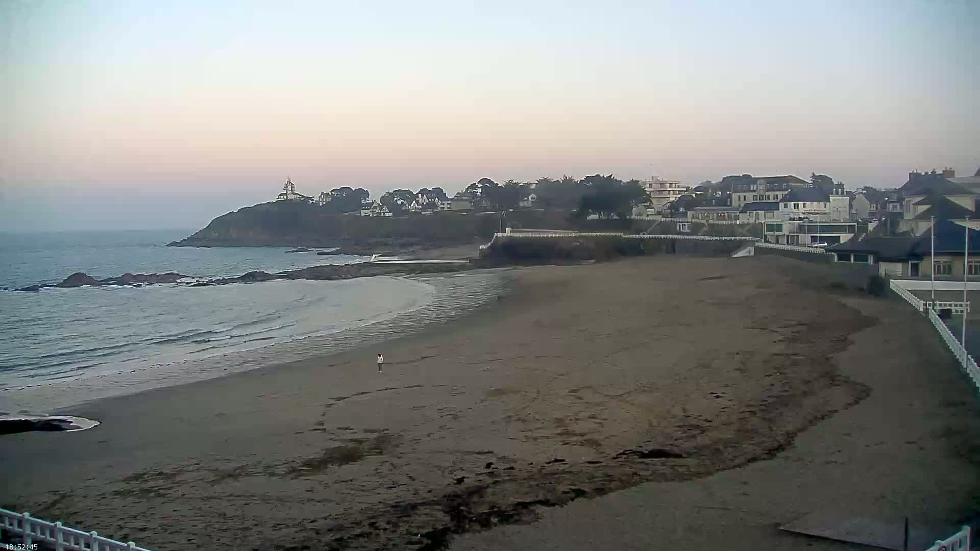 A sandy beach stretches out to a calm ocean under a pale, overcast sky, with a small town and a prominent headland visible in the background.