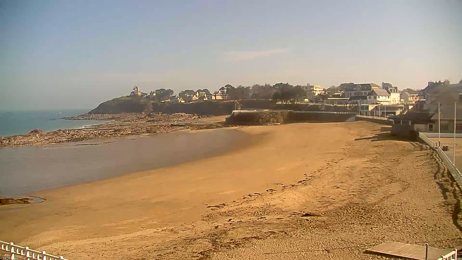 A sandy beach stretches out to a calm ocean, with a small town and a lighthouse visible on a cliff in the distance under a clear, sunny sky.
