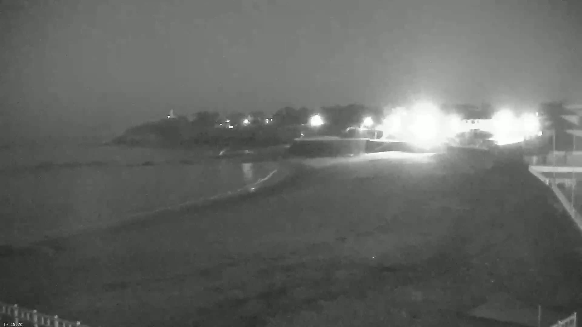 A nighttime, low-light image shows a dark beach and ocean with a dimly lit shoreline featuring buildings and bright lights.