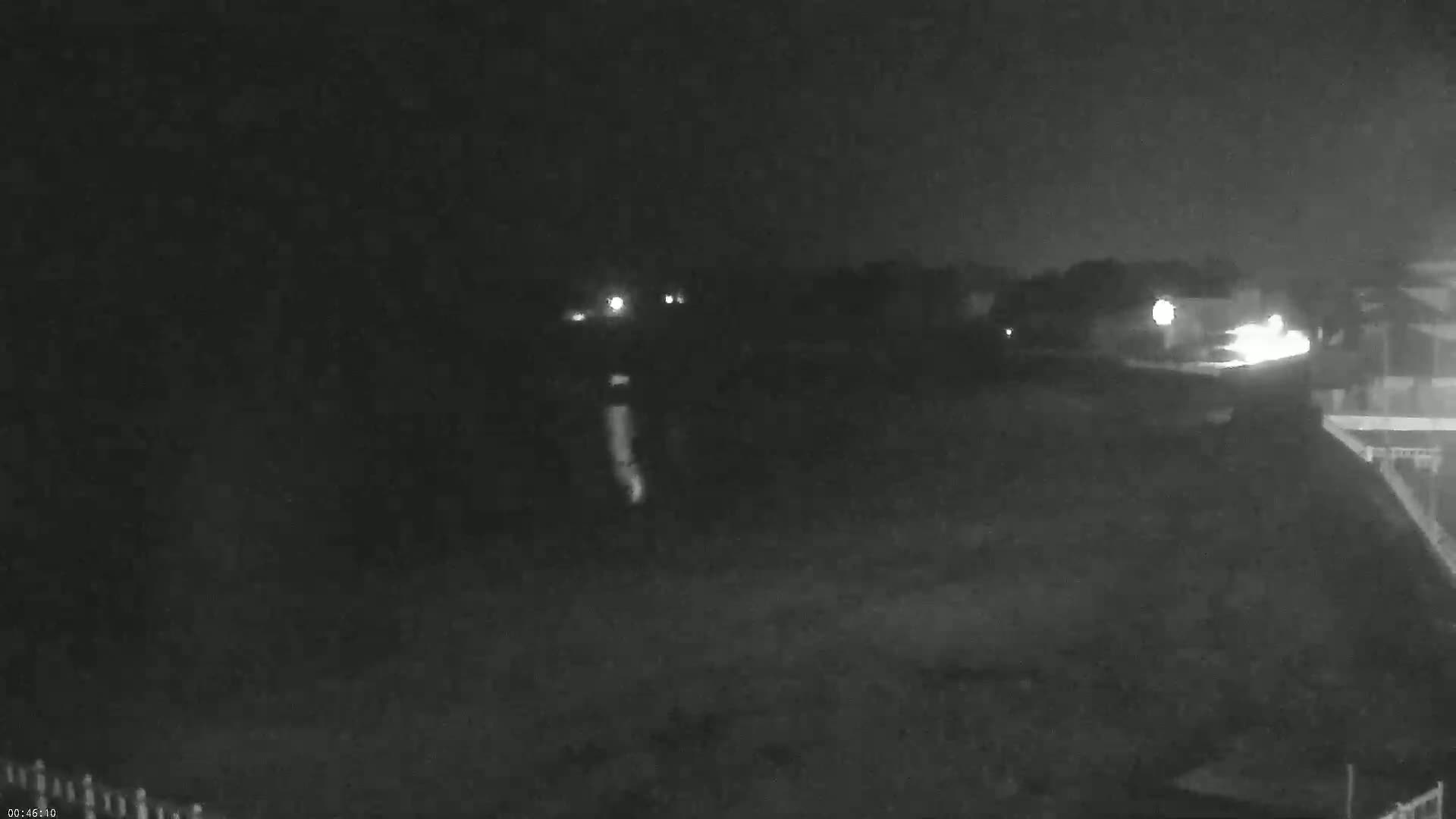 A nighttime, low-light view of a dark, mostly empty field with a few distant lights and a building on the right.