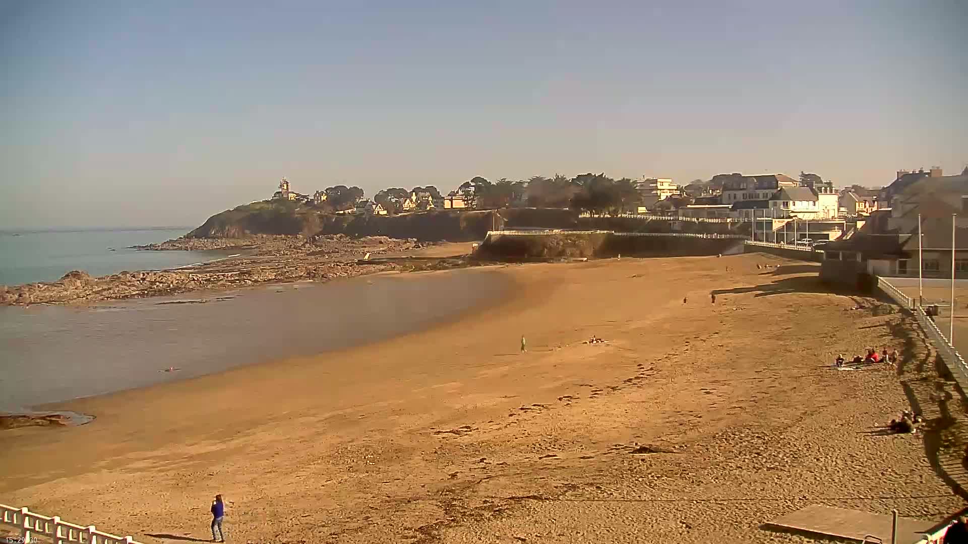 A sandy beach curves around a calm bay, with a small town and a lighthouse visible on a cliff in the background under a clear, sunny sky.
