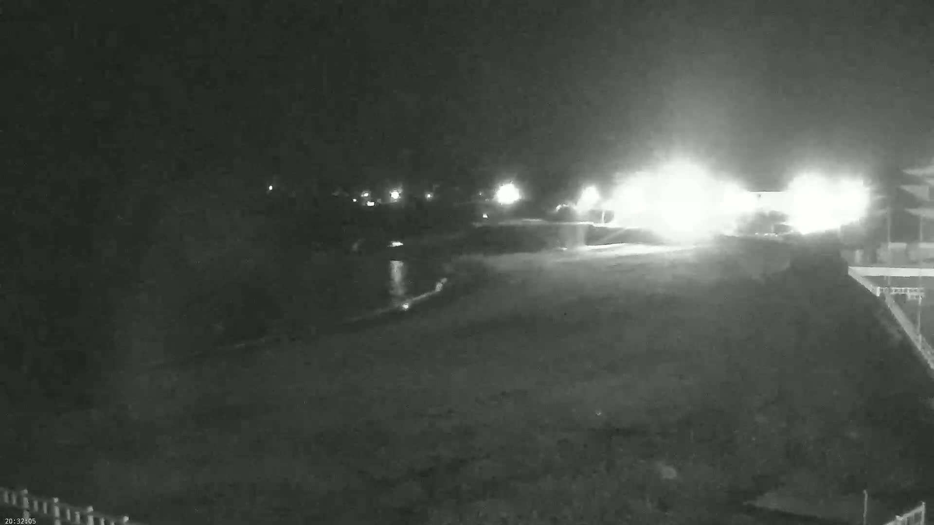 A nighttime, low-light view of a dark, seemingly empty expanse of land near a body of water, with bright lights visible in the distance.