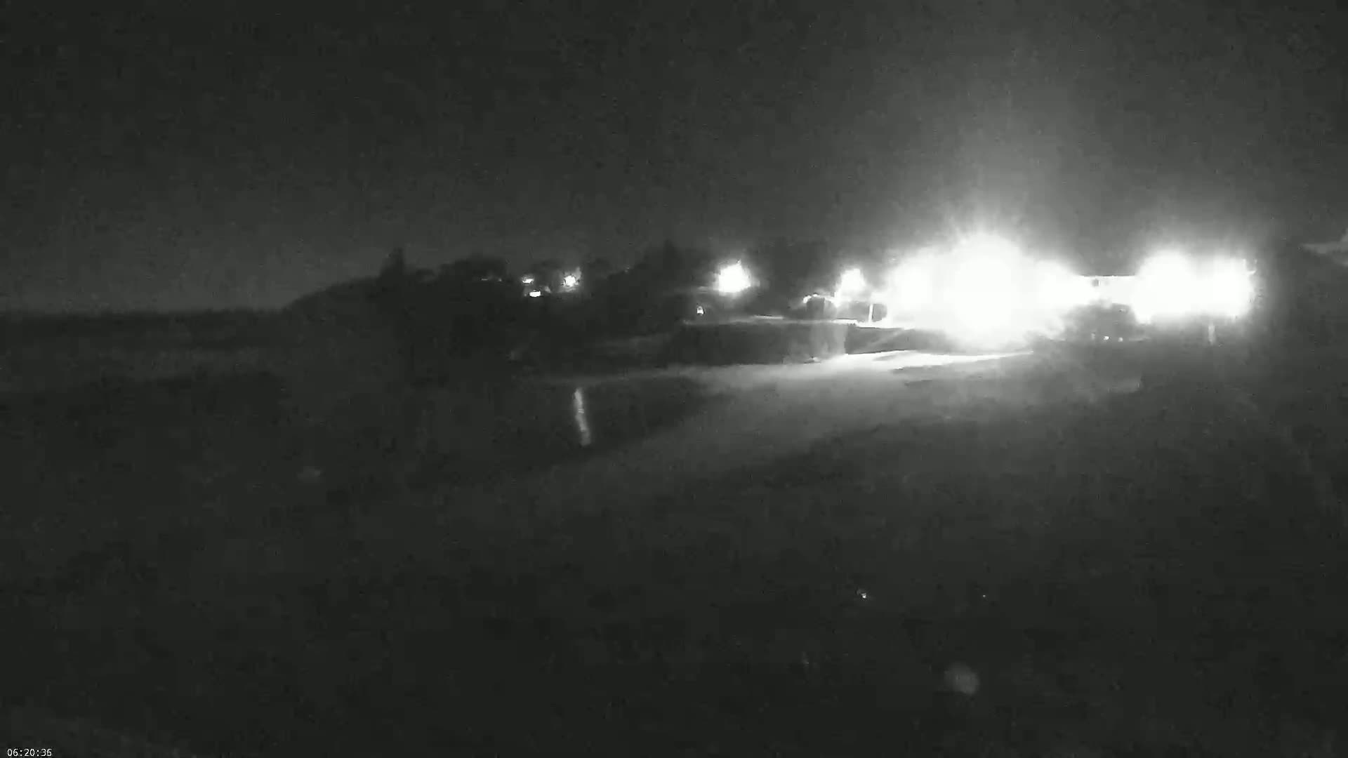 A nighttime, low-light view shows a road with bright headlights approaching from the distance, passing by some dark vegetation.