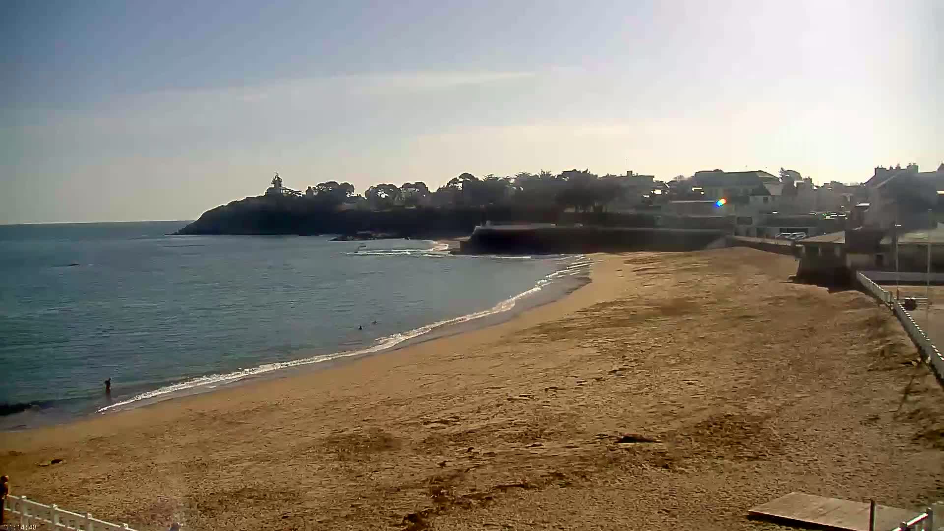 A sandy beach curves gently toward a calm ocean under a mostly sunny sky, with a small town and a distant lighthouse visible on a cliff in the background.