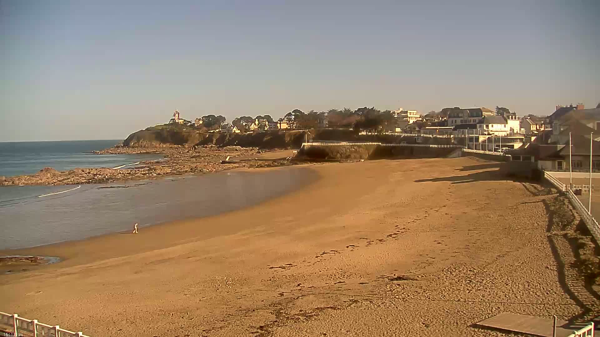 A sandy beach stretches out to a calm ocean under a clear, sunny sky, with a small town and a lighthouse visible on a cliff in the background.
