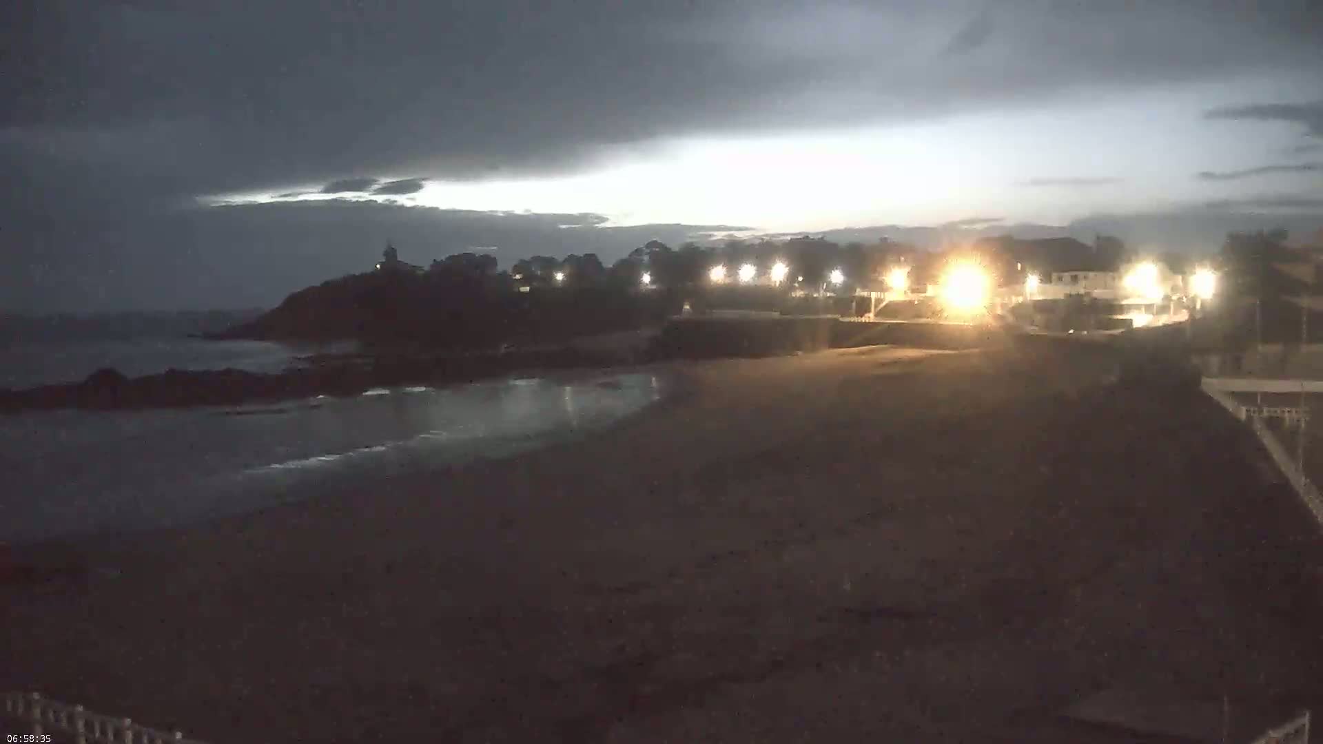 A dark, overcast night scene shows a dimly lit beach with a calm ocean, rocky shoreline, and buildings with lights in the background.