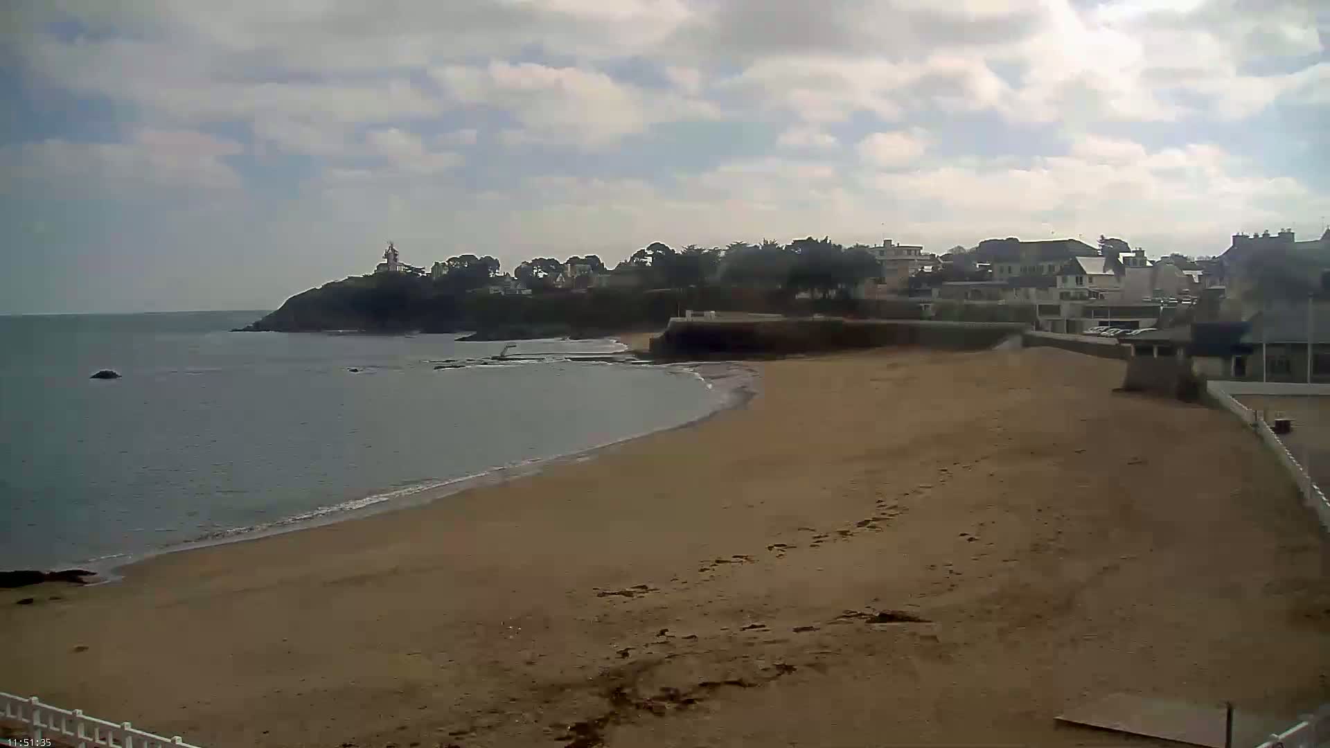 A sandy beach curves gently toward a calm sea under a partly cloudy sky, with a small town and a lighthouse visible on a distant headland.