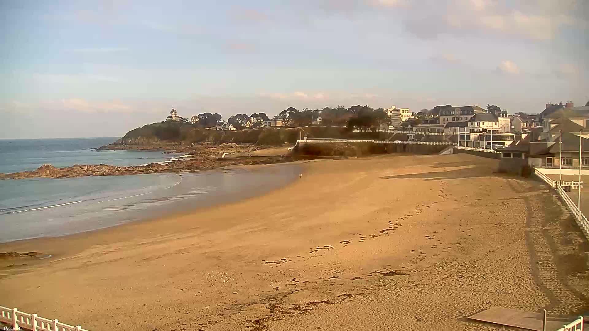 A sandy beach stretches towards a rocky shoreline and a village of buildings perched on a hill overlooking a calm ocean under a mostly clear sky.