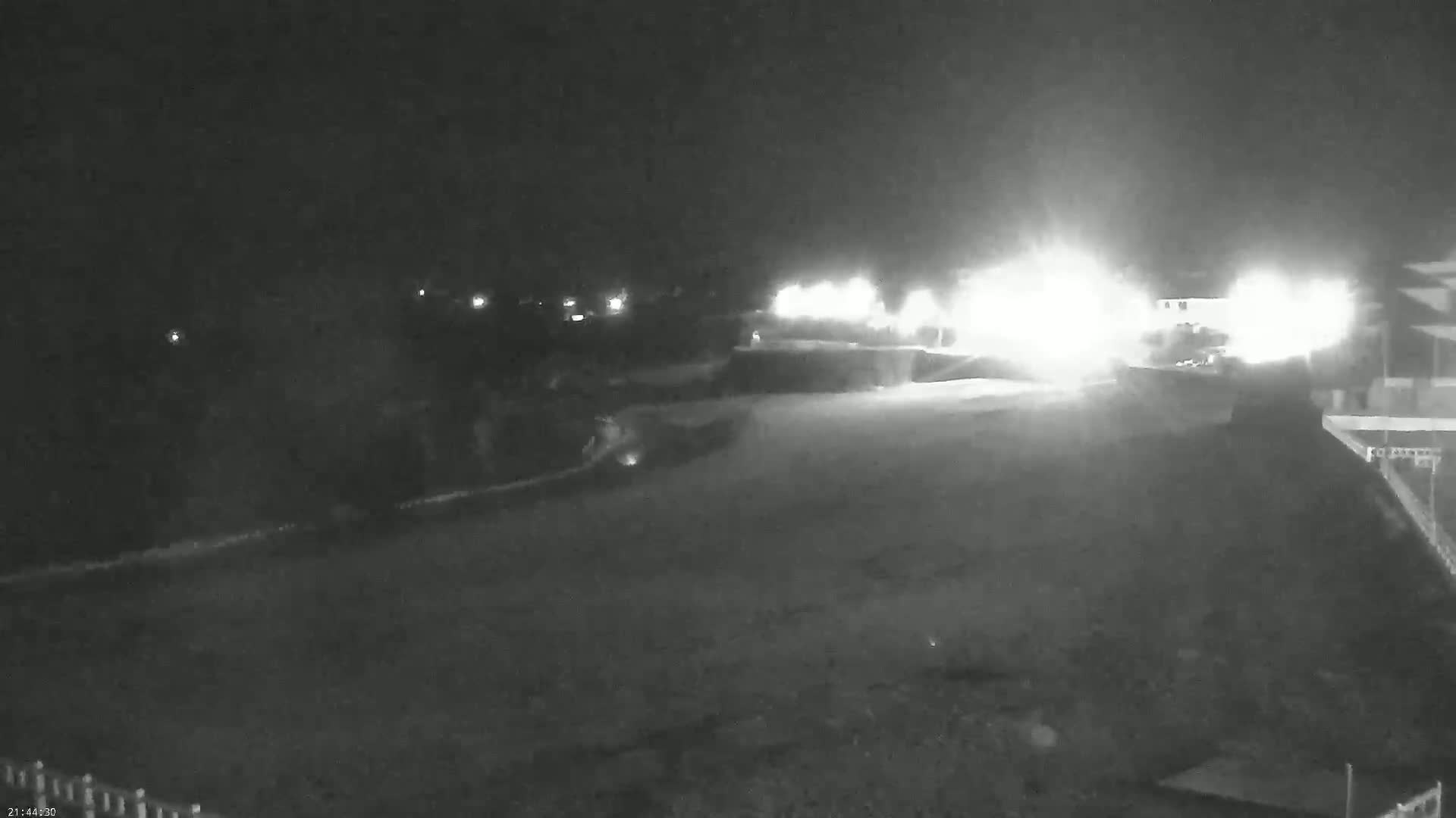 A nighttime, low-light view of a paved area with bright lights in the distance, possibly a roadway or parking lot.