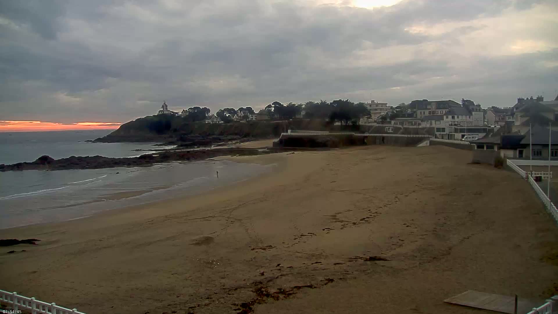 A sandy beach stretches out from a rocky coastline under a cloudy sky, with a town and a lighthouse visible on a hill in the distance, and a sunrise barely visible on the horizon.