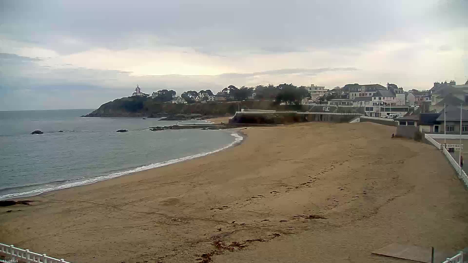 A sandy beach curves gently around a calm sea, toward a village perched atop a rocky cliff under an overcast sky.