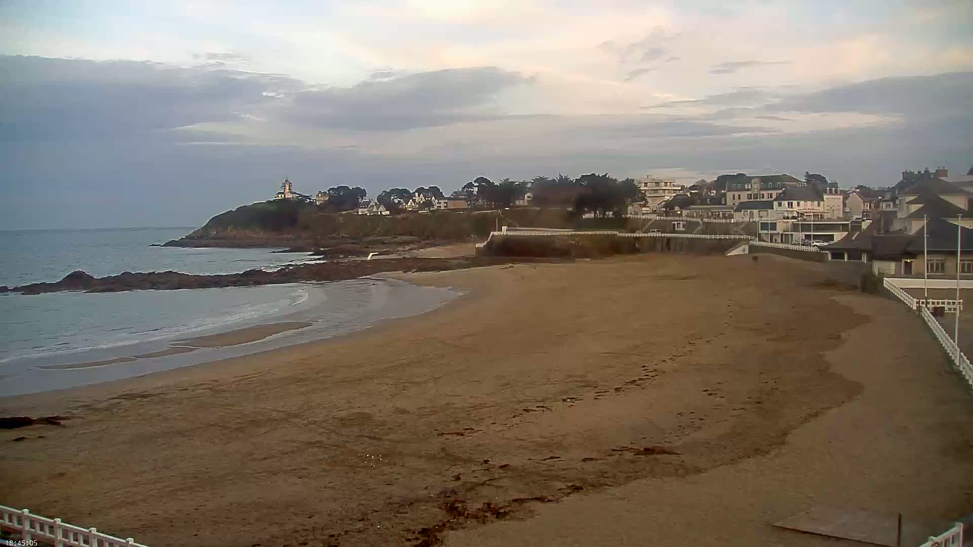 A sandy beach stretches towards a line of buildings perched on a rocky headland under a cloudy sky.