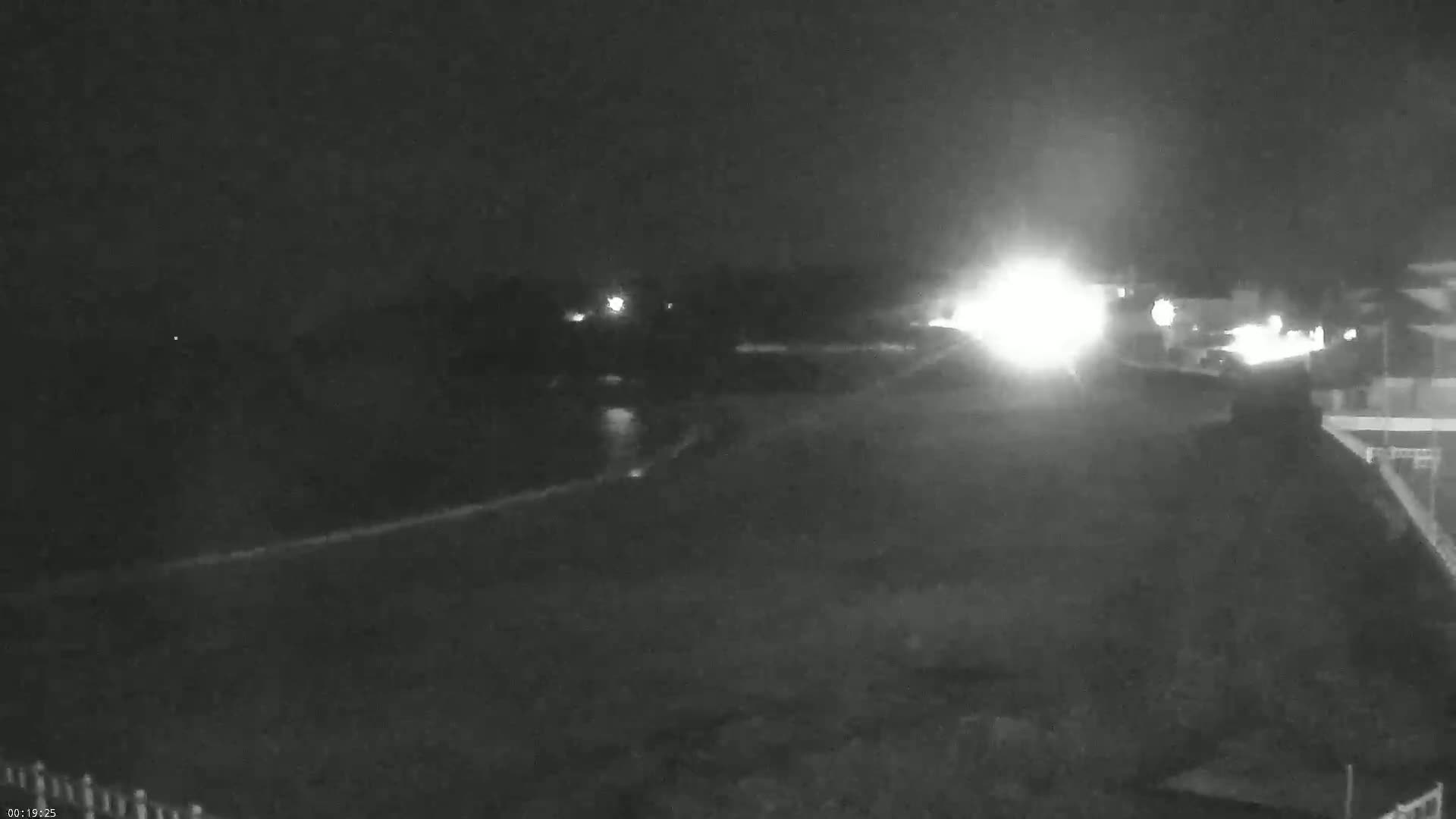 A nighttime, low-light view shows a dark, empty area with a bright light source in the distance, possibly from vehicle headlights, and some faint lights in the background.
