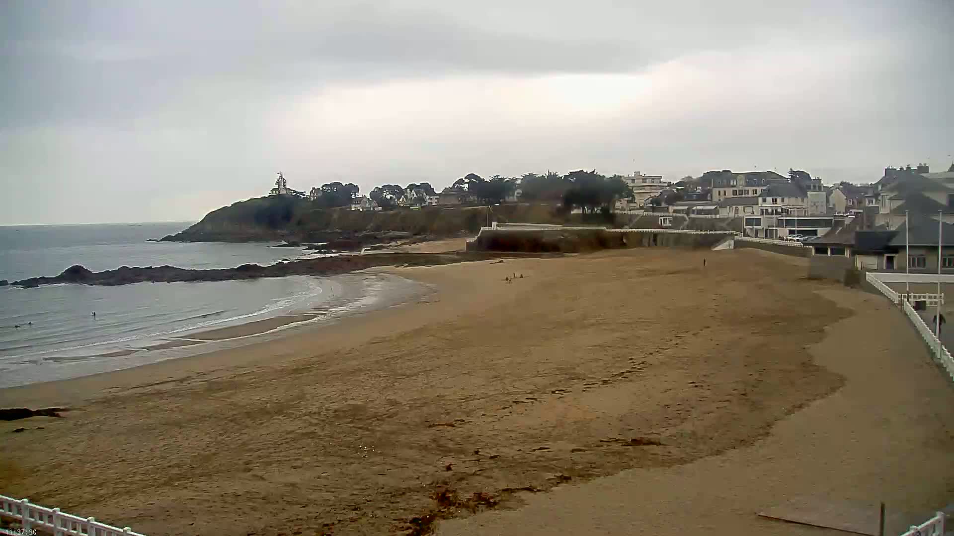 A sandy beach curves around a rocky coastline, under an overcast sky, with a small town and a lighthouse visible in the distance.