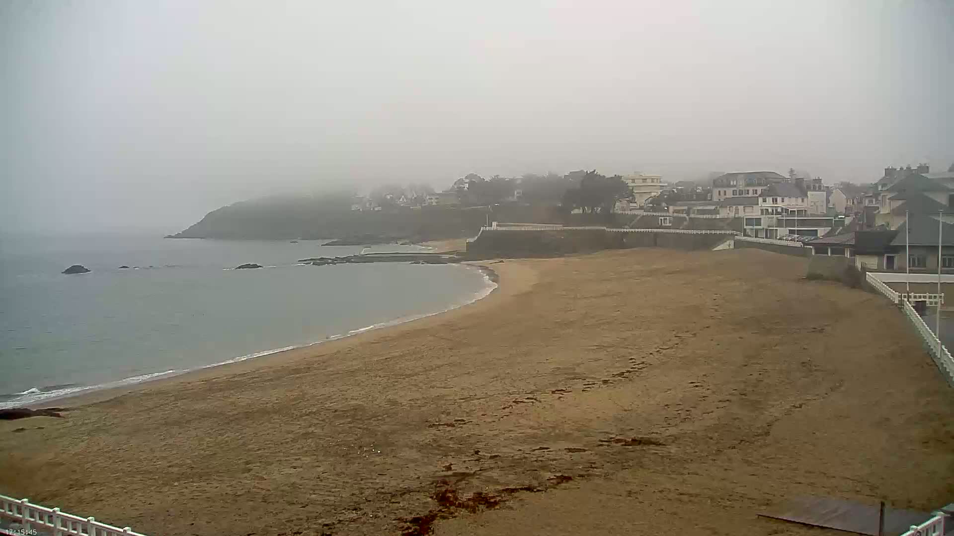 A foggy coastal scene shows a sandy beach curving around a calm bay towards a town nestled on a hillside.