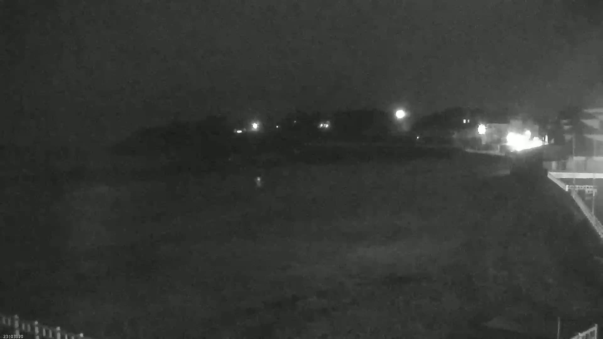 A nighttime, low-light view shows a dark expanse of land with a few distant lights, possibly a beach or field, under a clear, dark sky.