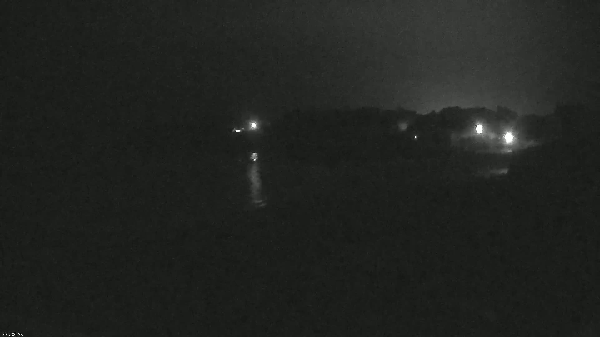 A dark, nighttime scene shows a body of water reflecting a few distant lights on the shore, under a foggy or hazy sky.