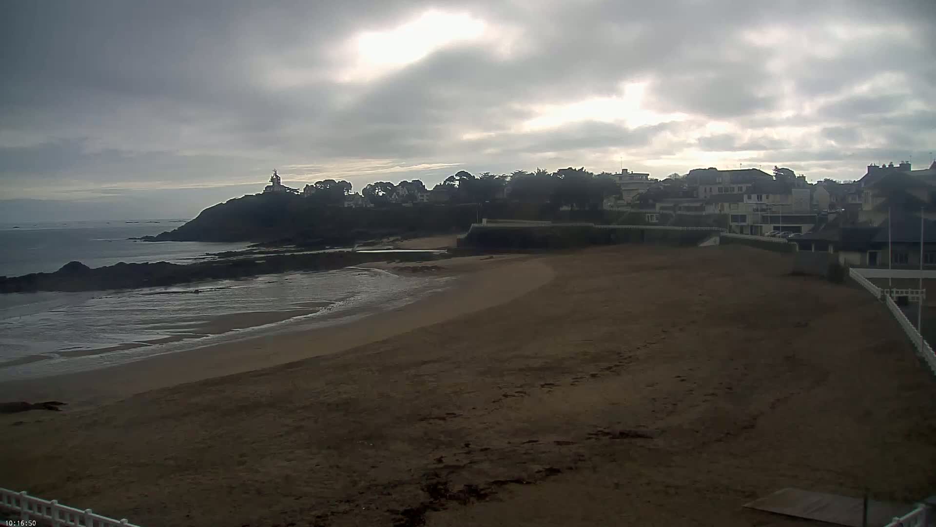 A sandy beach curves around a rocky shoreline, with a village and a tower visible on a hill under an overcast sky.