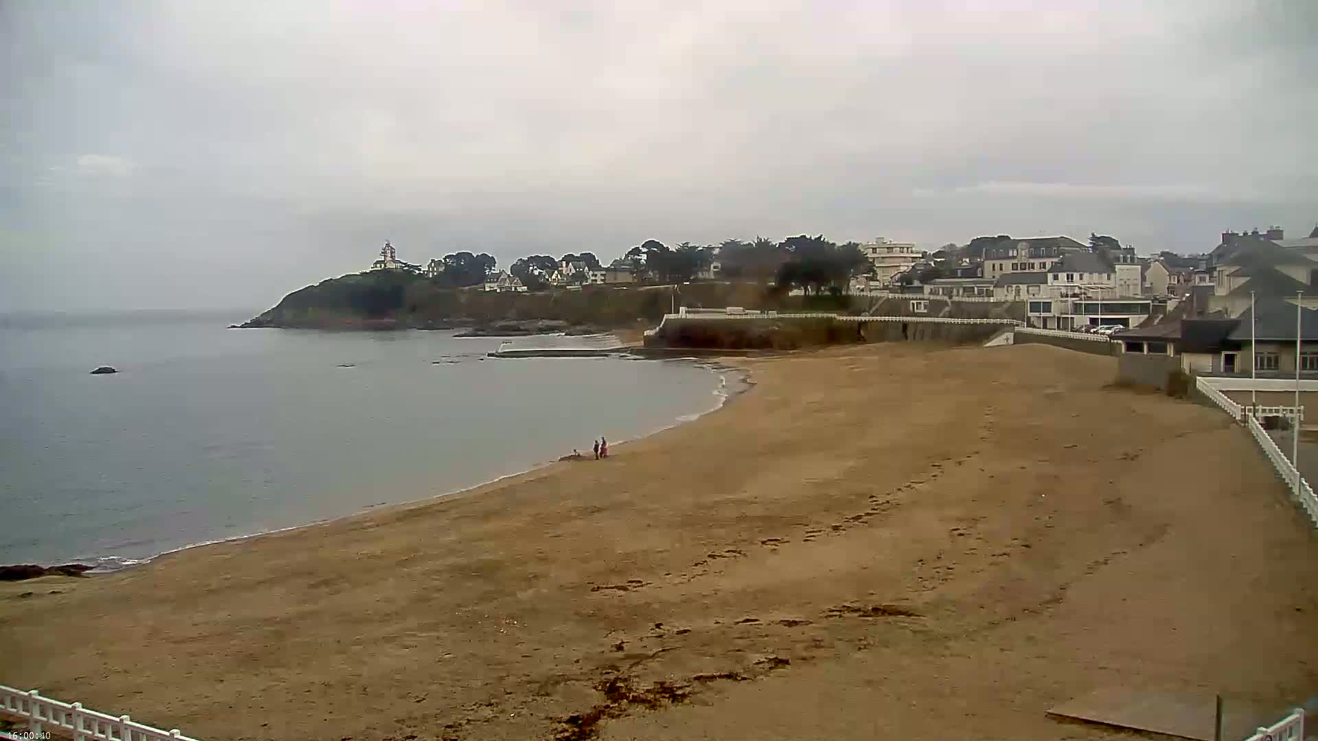 A sandy beach stretches out to a calm, grey sea under an overcast sky, with a small town and a lighthouse visible on a headland in the distance.