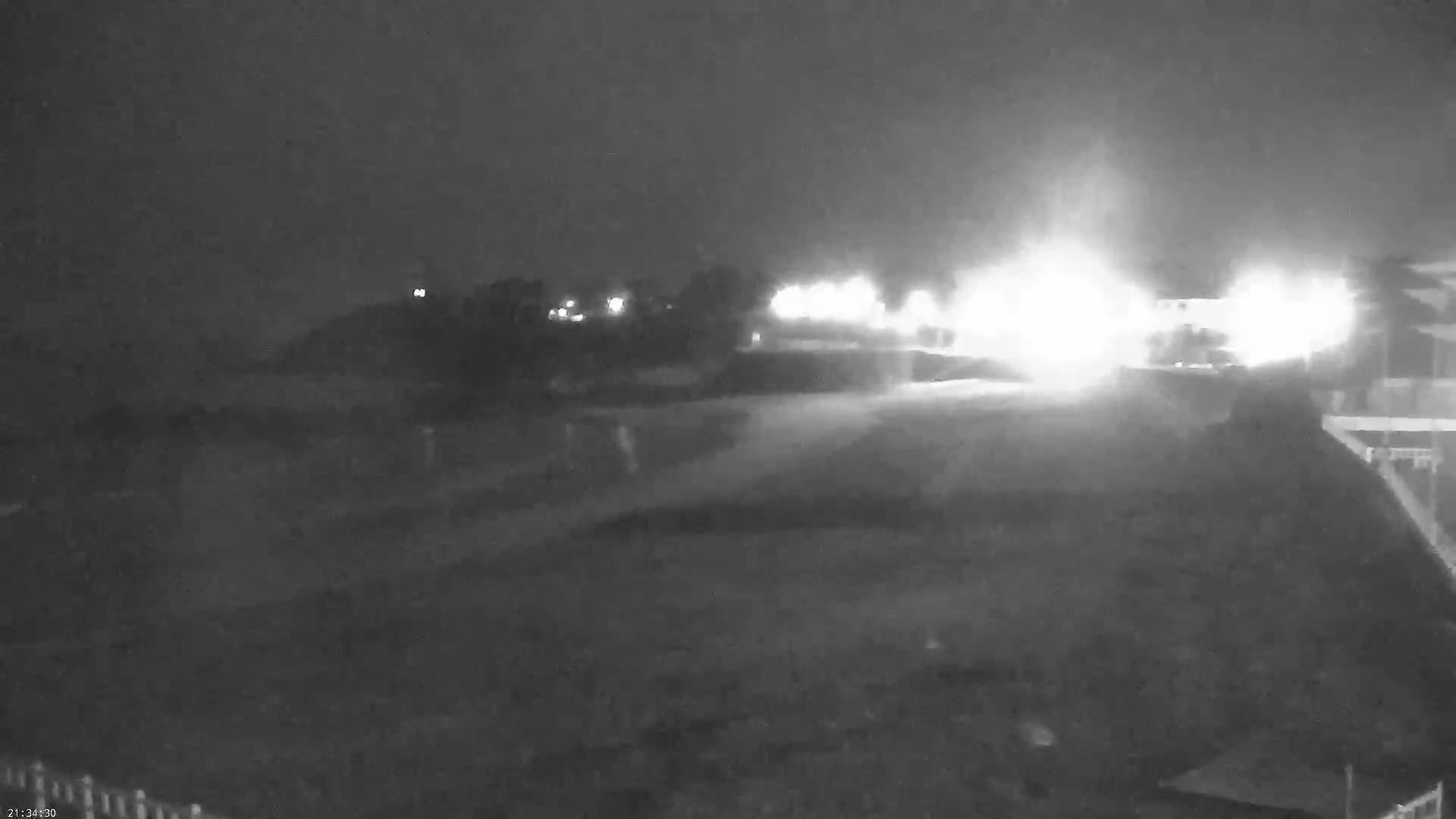 A nighttime, low-light view shows a dimly lit road with bright lights in the distance, surrounded by dark, possibly grassy areas.