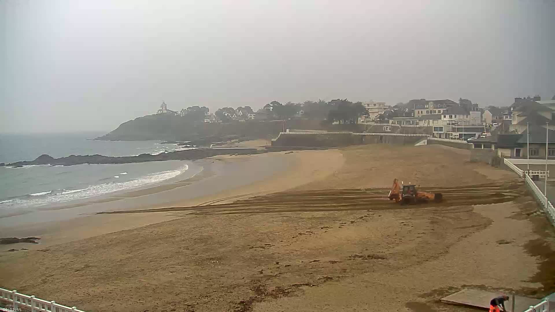 A bulldozer is working on a sandy beach next to a town on an overcast day, with a rocky coastline and ocean visible in the background.