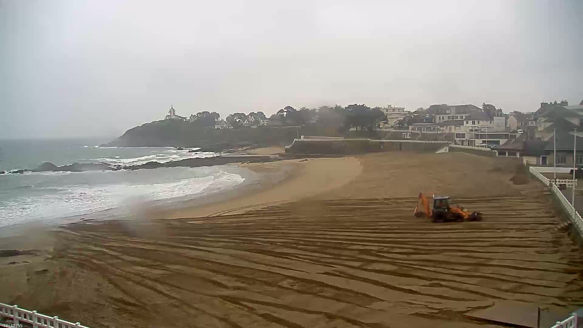 A sandy beach with a small orange piece of construction equipment is being worked on, next to a town on a cloudy day with waves rolling onto the shore.